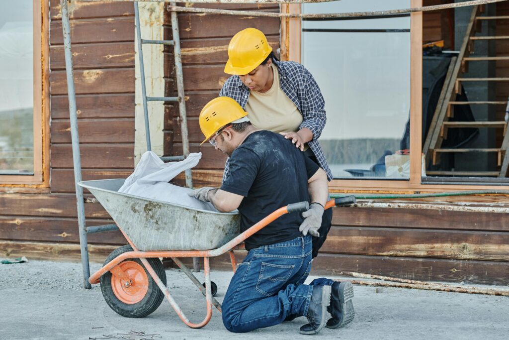 A construction worker receiving help while kneeling with a wheelbarrow on a job site.